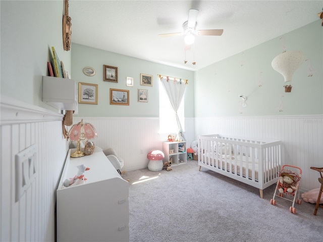 carpeted bedroom featuring wainscoting, ceiling fan, and a crib