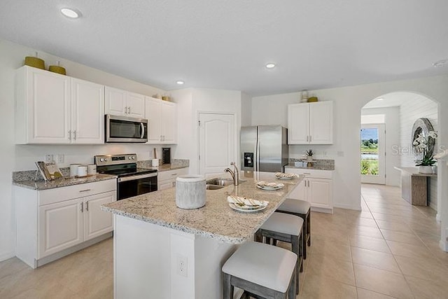 kitchen featuring arched walkways, appliances with stainless steel finishes, a sink, an island with sink, and a kitchen bar