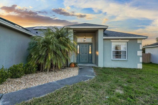 exterior entry at dusk featuring stucco siding, a shingled roof, and a yard