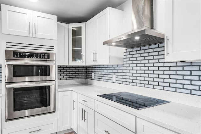 kitchen with white cabinets, black electric cooktop, wall chimney range hood, double oven, and backsplash