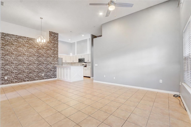 unfurnished living room featuring light tile patterned floors, ceiling fan with notable chandelier, brick wall, a high ceiling, and baseboards