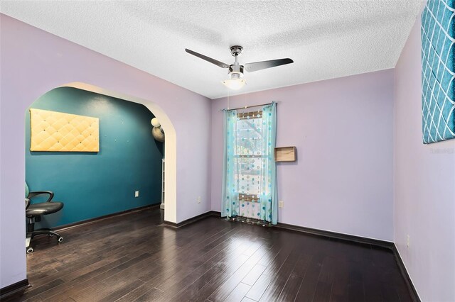 empty room with a textured ceiling, ceiling fan, arched walkways, wood finished floors, and baseboards