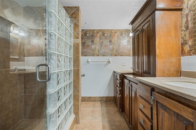 full bath featuring a textured ceiling, two vanities, a tile shower, wainscoting, and tile walls