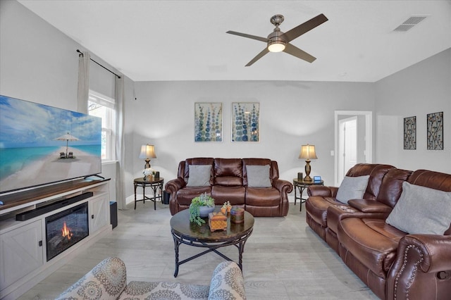 living room featuring light wood-style flooring, a glass covered fireplace, visible vents, and a ceiling fan