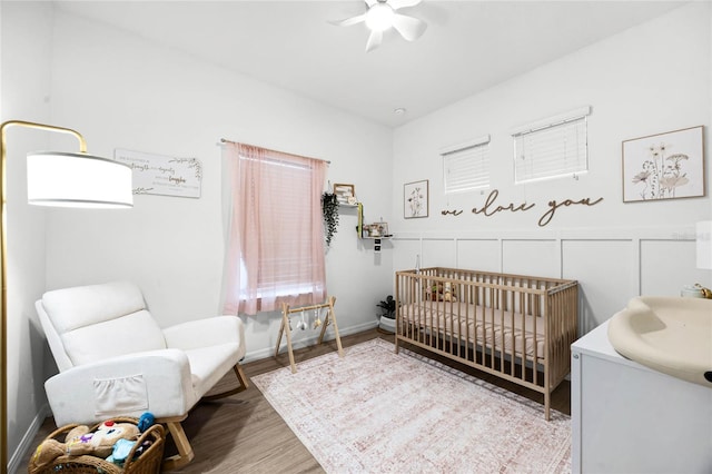 bedroom featuring a decorative wall, a ceiling fan, wainscoting, wood finished floors, and a crib