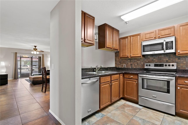 kitchen featuring stainless steel appliances, a sink, a ceiling fan, backsplash, and dark countertops