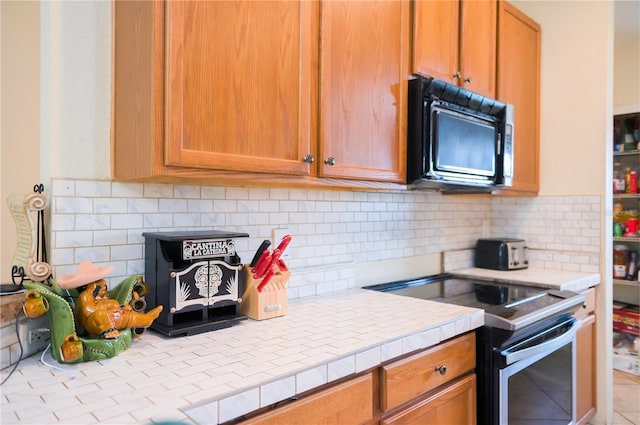 kitchen featuring black microwave, tile countertops, tasteful backsplash, brown cabinetry, and stainless steel range with electric stovetop