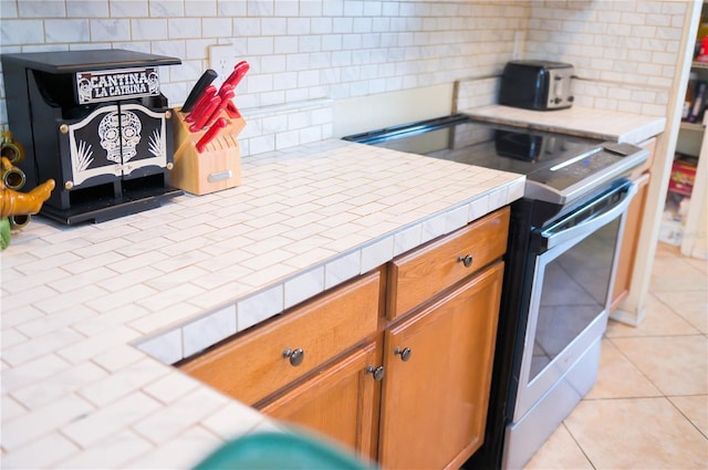 kitchen with light tile patterned floors, tasteful backsplash, tile counters, brown cabinets, and stainless steel electric stove