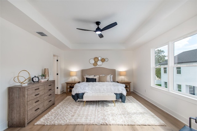 bedroom featuring a ceiling fan, light wood-style floors, visible vents, baseboards, and a tray ceiling