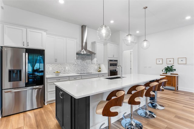 kitchen featuring light wood finished floors, decorative backsplash, appliances with stainless steel finishes, wall chimney range hood, and a sink