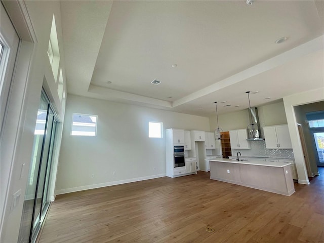 kitchen featuring light wood-style flooring, white cabinetry, wall chimney exhaust hood, a raised ceiling, and a large island with sink