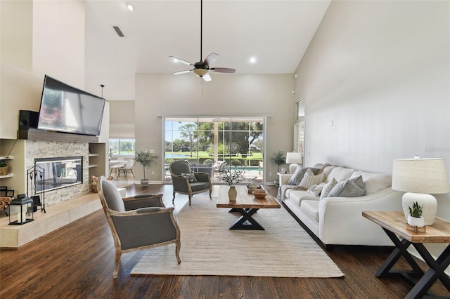 living room featuring baseboards, a towering ceiling, ceiling fan, wood finished floors, and a stone fireplace