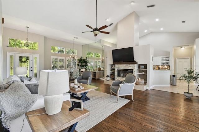 living room with visible vents, wood finished floors, a stone fireplace, high vaulted ceiling, and ceiling fan with notable chandelier