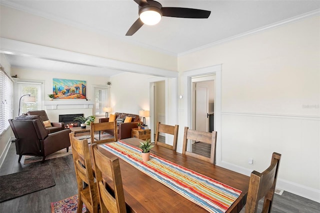 dining space with a warm lit fireplace, dark wood-style floors, ceiling fan, a wainscoted wall, and crown molding