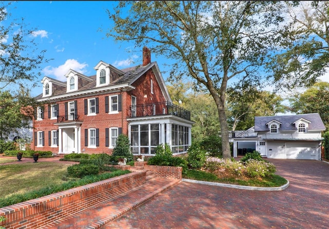 colonial house featuring a balcony, a garage, brick siding, a sunroom, and a chimney