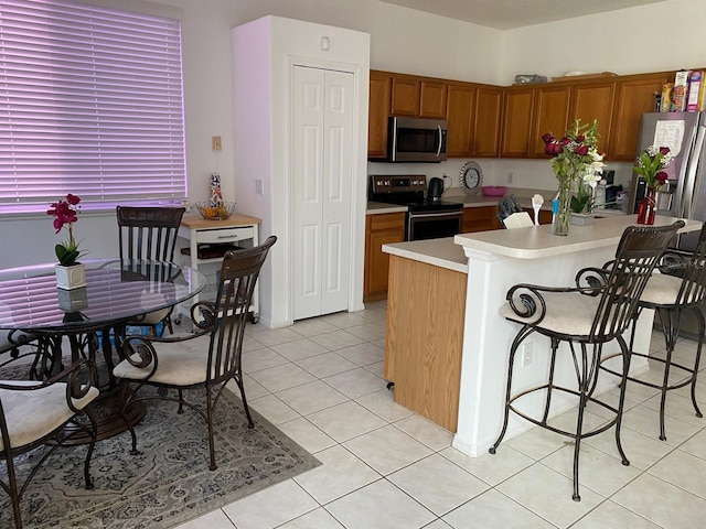 kitchen with light tile patterned floors, brown cabinetry, a breakfast bar area, stainless steel appliances, and light countertops