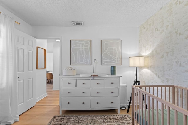 bedroom featuring visible vents, a crib, a textured ceiling, light wood-style floors, and wallpapered walls