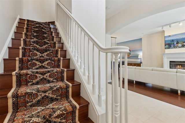 stairs with arched walkways, wood finished floors, a glass covered fireplace, and rail lighting