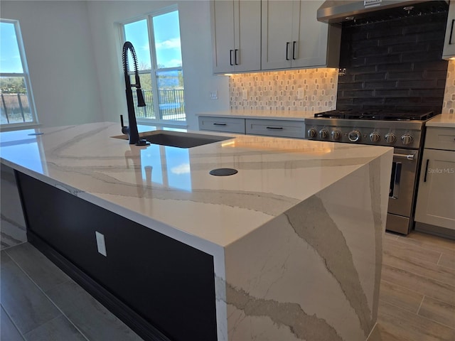 kitchen featuring stainless steel stove, backsplash, a sink, wall chimney range hood, and light stone countertops