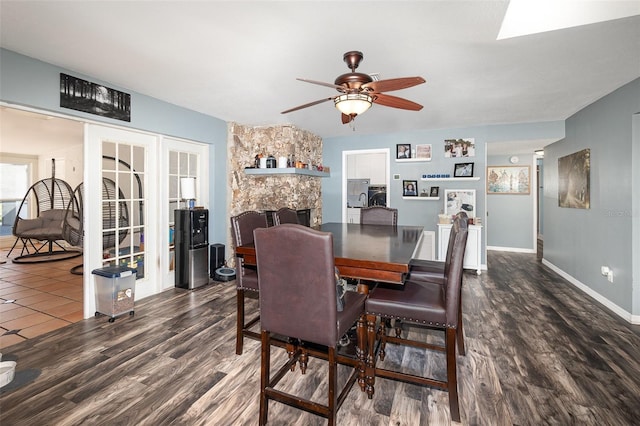 dining area with ceiling fan, a stone fireplace, a skylight, baseboards, and dark wood finished floors