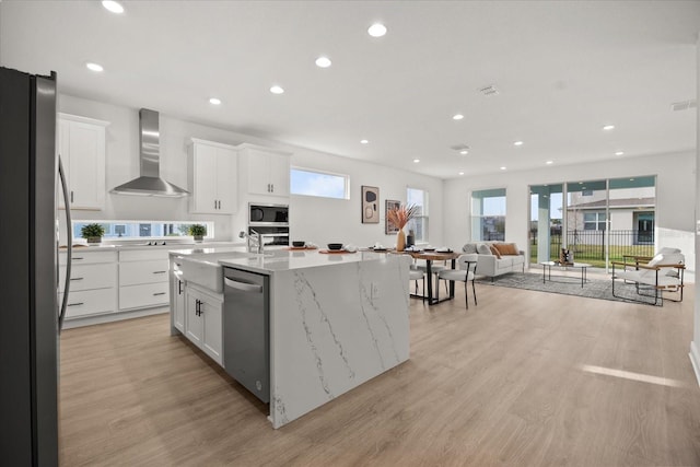 kitchen with light wood finished floors, stainless steel appliances, white cabinetry, a sink, and wall chimney range hood