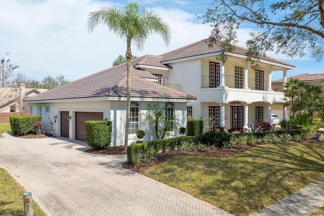 view of front of home featuring an attached garage, a balcony, decorative driveway, stucco siding, and a front lawn