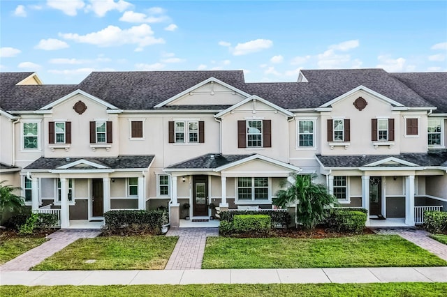 townhome / multi-family property with a shingled roof, covered porch, a front lawn, and stucco siding