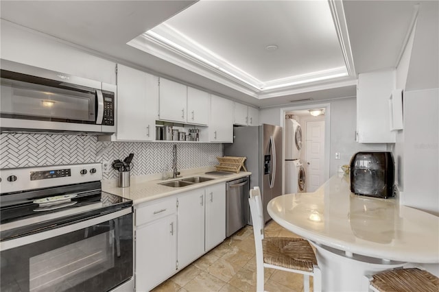 kitchen with a tray ceiling, stainless steel appliances, light countertops, stacked washing maching and dryer, and a sink