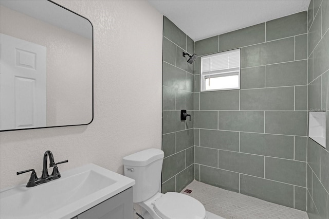 bathroom with a textured wall, tiled shower, vanity, and toilet