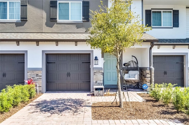 entrance to property featuring decorative driveway, stucco siding, a shingled roof, an attached garage, and stone siding