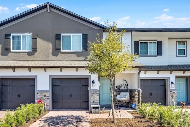 townhome / multi-family property featuring driveway, stone siding, roof with shingles, an attached garage, and stucco siding