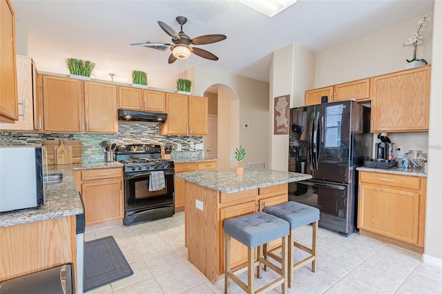 kitchen with arched walkways, a kitchen island, light stone counters, under cabinet range hood, and black appliances