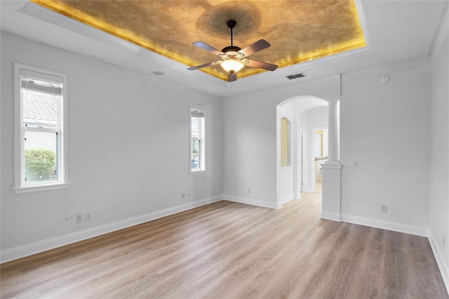 empty room with decorative columns, arched walkways, baseboards, light wood-style flooring, and a tray ceiling