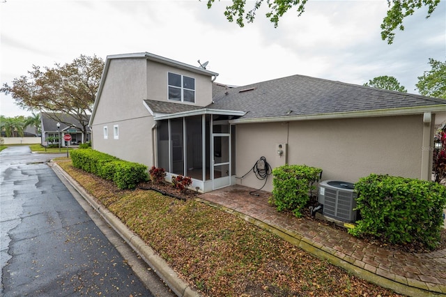 rear view of property with stucco siding, central air condition unit, a sunroom, and roof with shingles