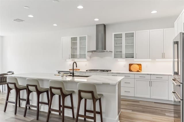 kitchen featuring white cabinets, an island with sink, wall chimney exhaust hood, fridge, and stainless steel gas stovetop