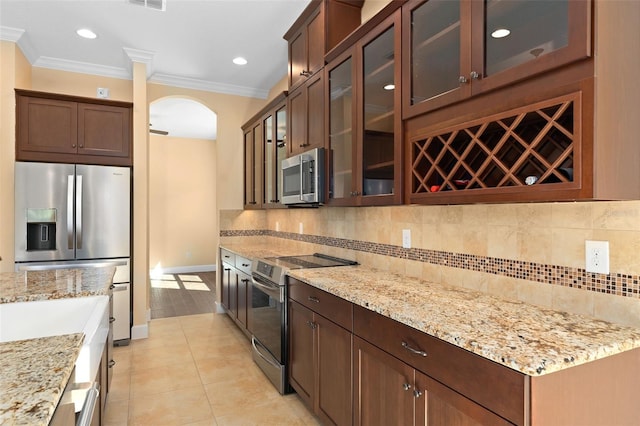 kitchen featuring light stone counters, arched walkways, backsplash, appliances with stainless steel finishes, and ornamental molding