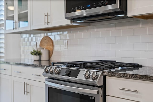 kitchen featuring stainless steel appliances, white cabinets, decorative backsplash, dark stone countertops, and glass insert cabinets