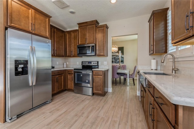 kitchen featuring a textured ceiling, a notable chandelier, stainless steel appliances, light wood finished floors, and brown cabinetry