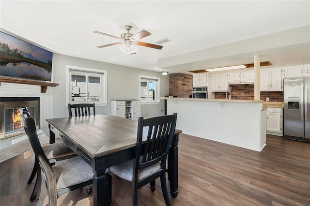 dining space with a brick fireplace, visible vents, dark wood-style floors, and ceiling fan