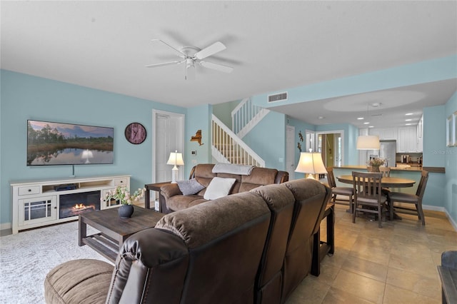 living room featuring visible vents, a glass covered fireplace, ceiling fan, baseboards, and stairs