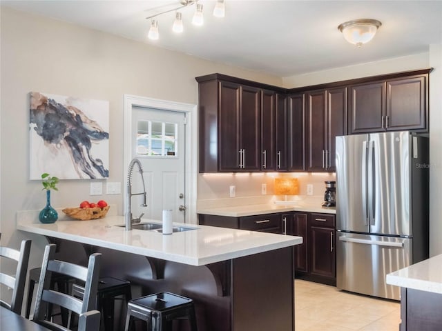 kitchen with light countertops, freestanding refrigerator, a sink, dark brown cabinetry, and a kitchen breakfast bar