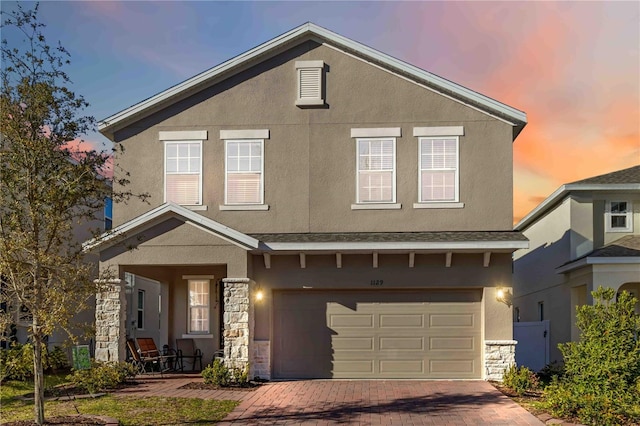 traditional-style house with a garage, stone siding, decorative driveway, and stucco siding