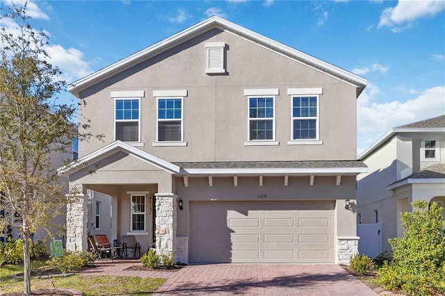 traditional-style house with a garage, stone siding, decorative driveway, and stucco siding