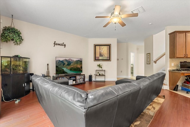 living room featuring light wood finished floors, visible vents, baseboards, ceiling fan, and stairs