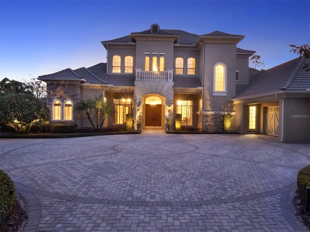 mediterranean / spanish-style home featuring decorative driveway, a tile roof, stucco siding, a balcony, and stone siding