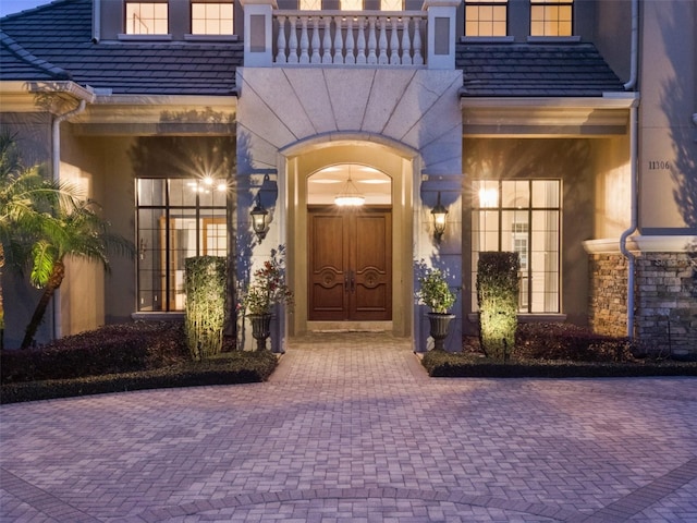 view of exterior entry with a balcony, stone siding, a high end roof, and stucco siding