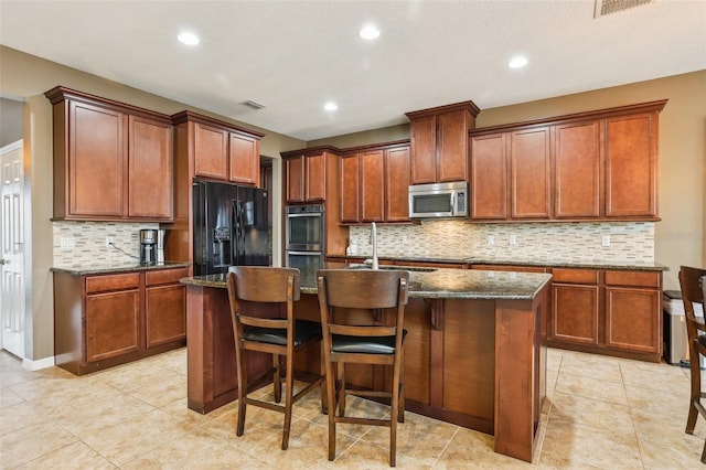 kitchen with stainless steel appliances, visible vents, a sink, an island with sink, and dark stone counters