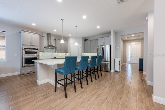 kitchen with stainless steel appliances, gray cabinetry, decorative backsplash, light wood-type flooring, and wall chimney exhaust hood