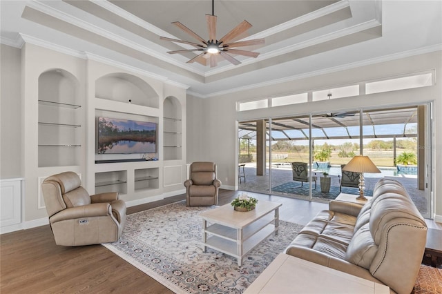 living area with a ceiling fan, a sunroom, wood finished floors, a tray ceiling, and built in shelves