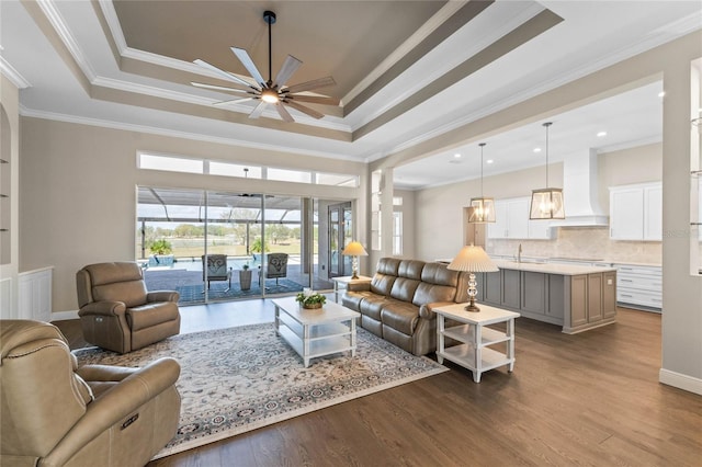 living room with dark wood-type flooring, a raised ceiling, crown molding, and a ceiling fan